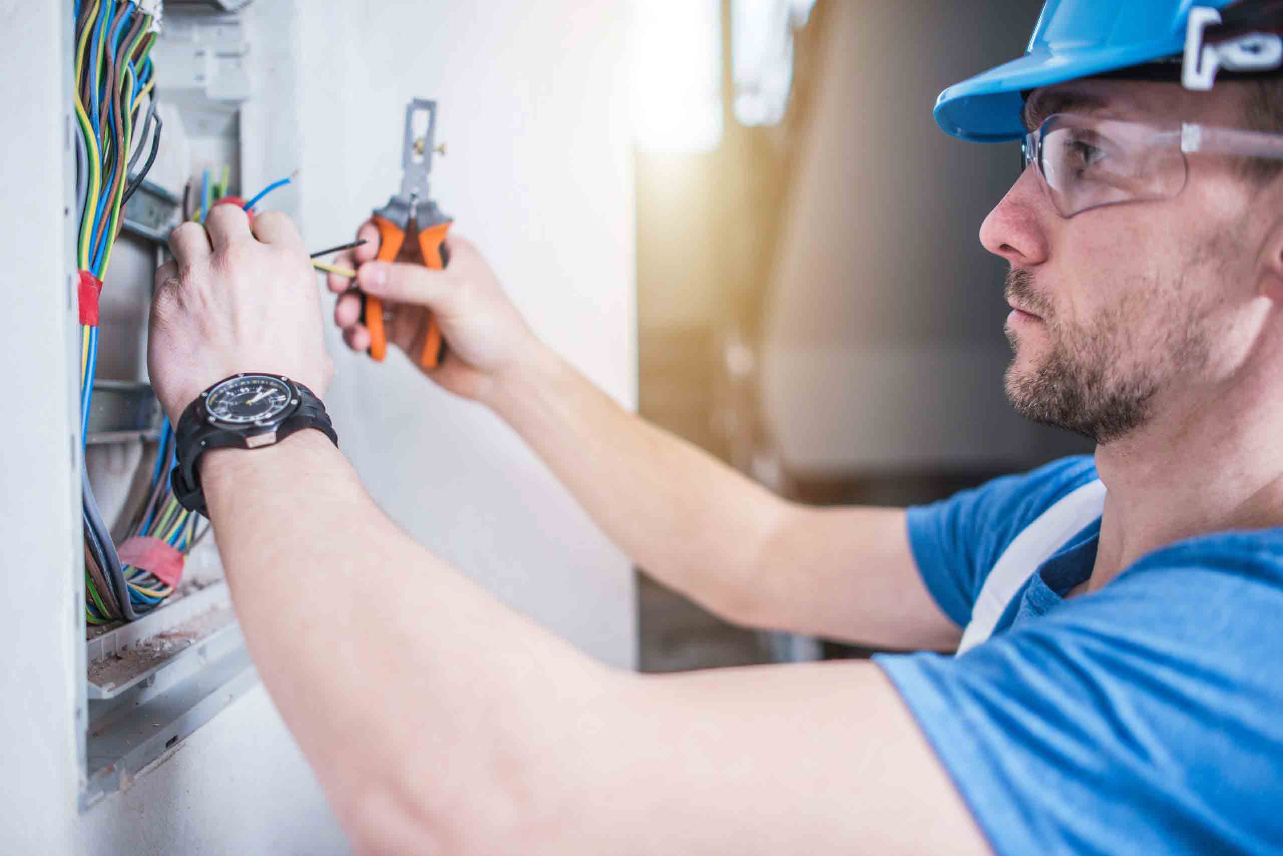 Male electrician wearing goggles and hard hat, using wire cutters on colored wiring for home surge protection installation.