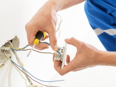 Person replacing a damaged electrical outlet, using a screwdriver, with exposed wiring in the background, emphasizing electrical safety and maintenance.