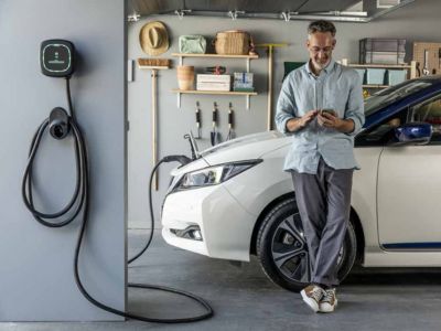Man using smartphone next to an electric vehicle charging station in a garage, highlighting the convenience of at-home EV charging for homeowners.