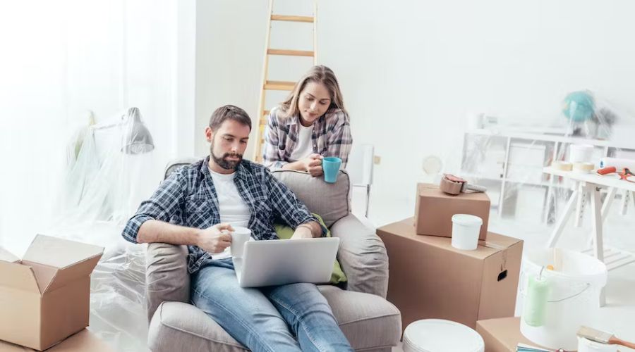 Couple in a partially renovated living room, sitting on a couch with a laptop, discussing home improvement ideas amidst moving boxes and paint supplies, reflecting on electrical upgrades for 2024.