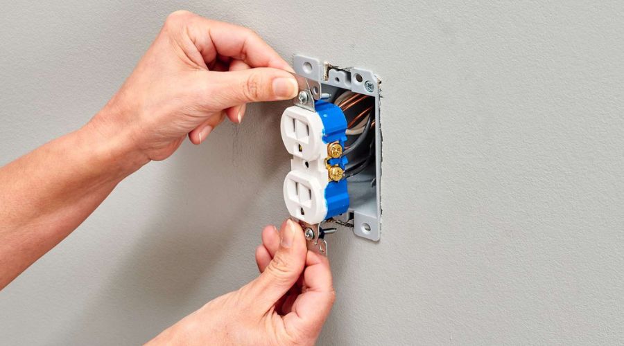 Hands installing a new electrical outlet with blue wiring components against a neutral wall, illustrating the importance of outlet replacement for electrical safety in homes.