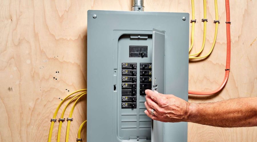 Hand opening electrical panel with circuit breakers, surrounded by yellow and orange wiring, illustrating signs for electrical panel replacement and upgrades.
