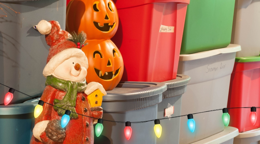 Snowman figurine in Santa attire with Halloween pumpkins, colorful lights, and storage bins, representing holiday decor.