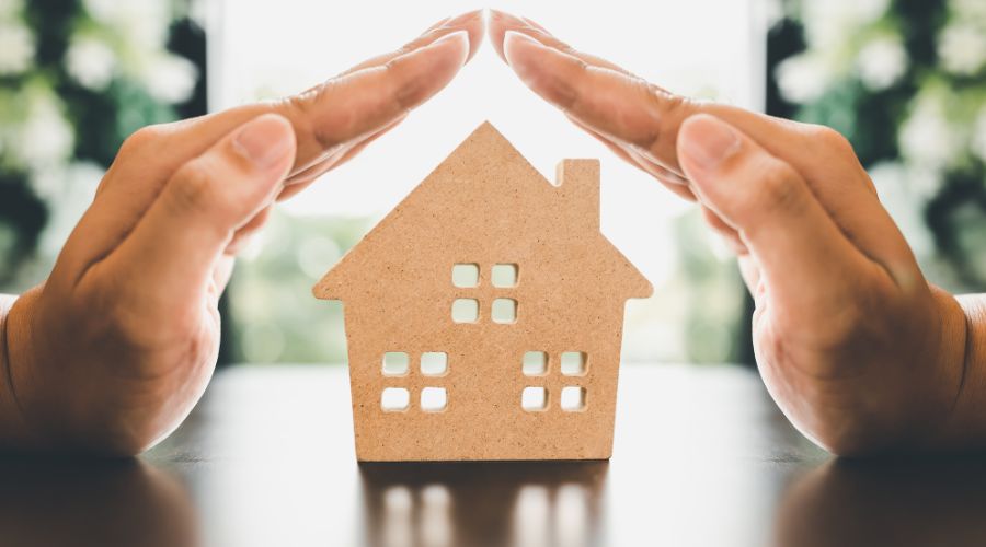 Hands protecting a small house model, symbolizing safety and security for homes and families, relevant to electrical safety measures and upgrades.