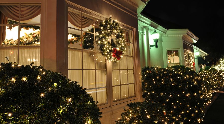 Holiday-decorated home exterior featuring a festive wreath with red bow, illuminated windows, and sparkling lights on surrounding bushes, emphasizing safe holiday decorating practices.